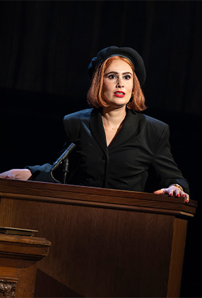 A performer in a vibrant costume poses dramatically on stage, with colourful lighting and a dark background.