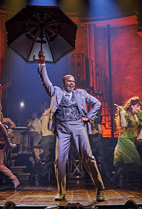 Cedric Neal performs on stage in Hadestown, wearing a dark suit, with dramatic lighting and a textured backdrop.