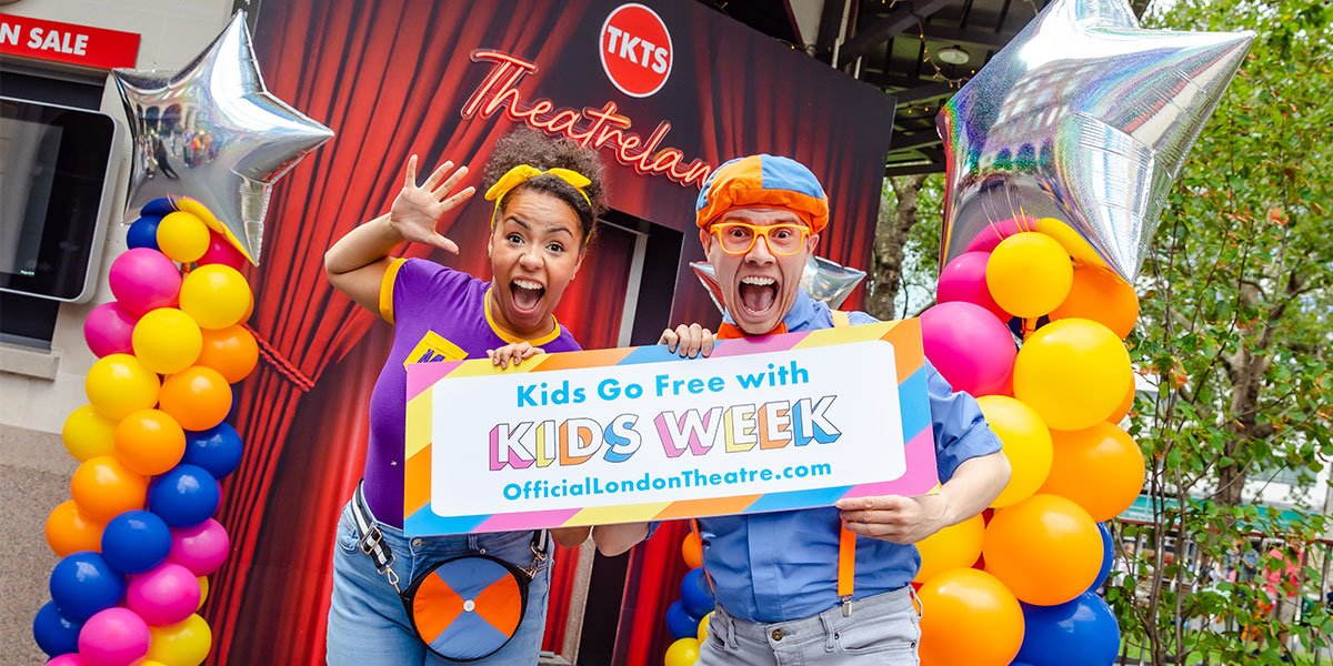 Two people dressed in colourful outfits hold a sign saying 'Kids go free with Kids Week'
