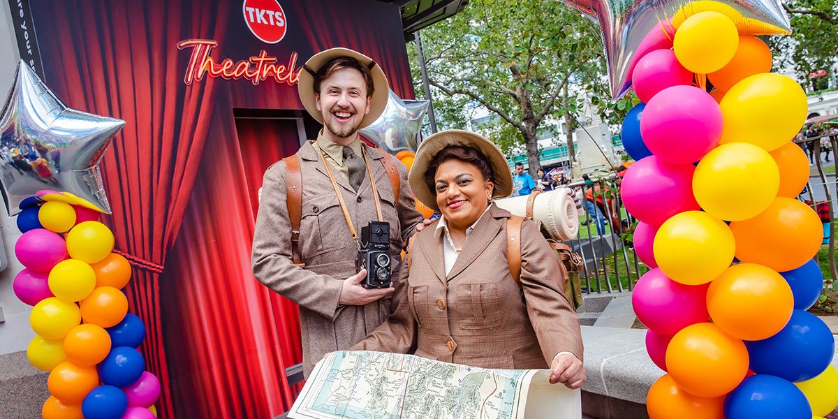 Two people stand wearing safari style outfits holding a camera and holding a map