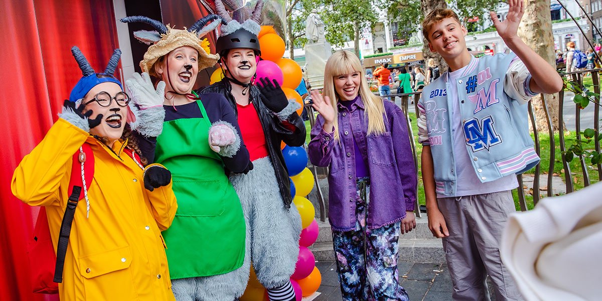 Three people dressed as goats stand with kids who are waving