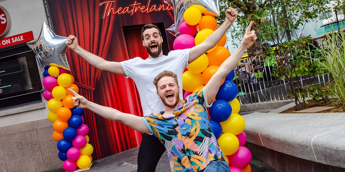 Two people stand with their arms outstretched, one wearing a white tshirt, one wearing a colourful shirt