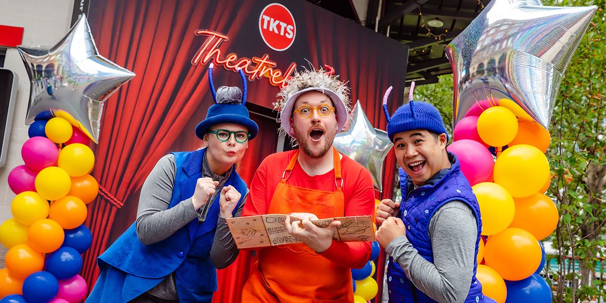 3 people stand, 1 in the centre in orange and 1 either side in blue with hats