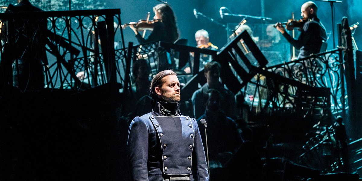 Bradley Jaden stands confidently in a dark uniform, with musicians visible in the background.