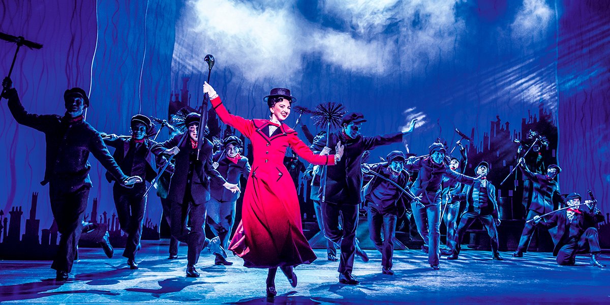 Zizi Strallen in a vibrant red dress, performing with dancers in dark costumes.
