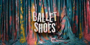 The image features an artistic backdrop made of flowing, draped fabric in shades of teal, pink, yellow, and hints of white. The fabric appears to be splattered with colors. In the center of the image, large, bold white text reads "BALLET SHOES". It is hung by rope as if a wooden sign. The letters are slightly worn and splattered with colorful paint. There is a used paintbrush on the floor.