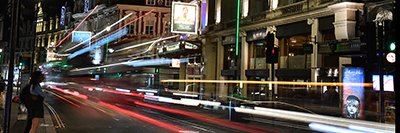 A busy theatre street at night with blurred lights and moving traffic creating vibrant motion.