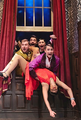 A group of actors in colourful costumes performs a comedic scene on a theatre stage, with props.