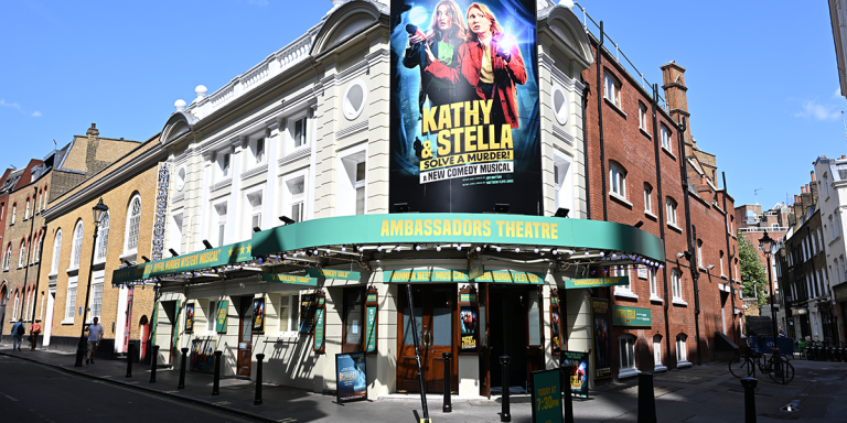 Kathy and Stella stand on stage, dressed in vibrant costumes, with a theatre backdrop behind them.