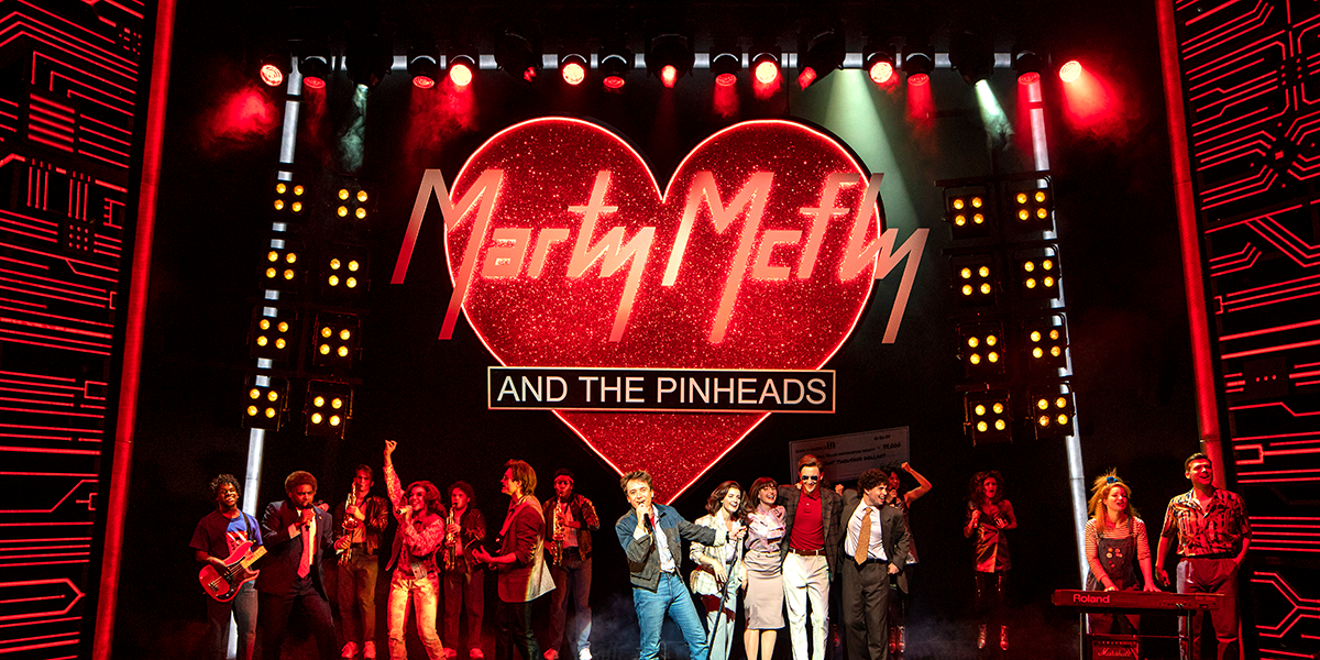 The cast of Back to the Future - The Musical poses together, smiling, in colourful costumes on stage.